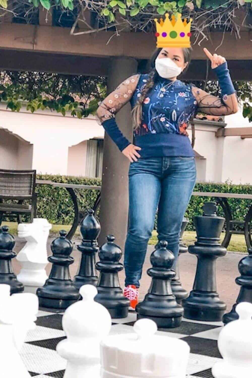 Girl standing on life size chess board in blue sweater and jeans with COVID-19 N95 mask pointing to a yellow emoji queen's crown on her head at the Hilton Santa Barbara