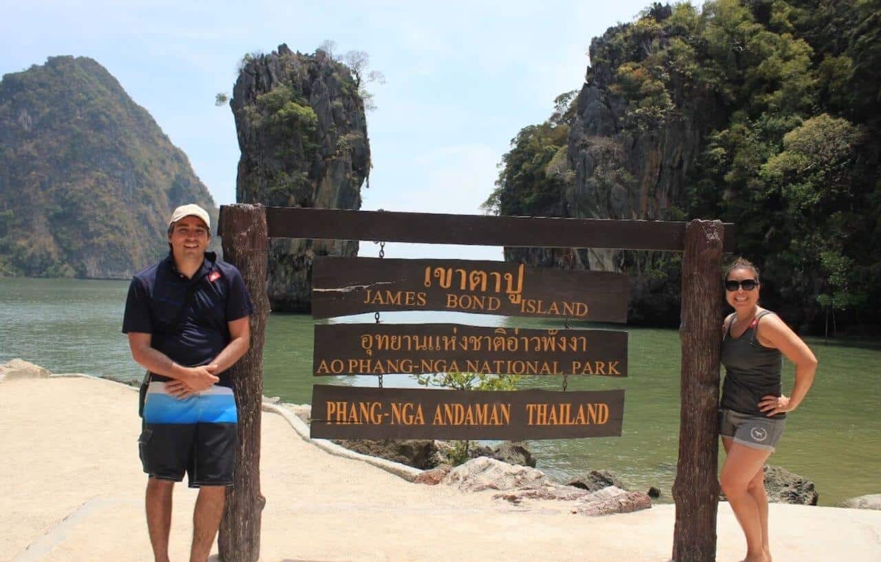 Boy and girl standing on either side of sign reading James Bond Island in English and Thai with ocean water and grass covered mountains in Phuket Thailand