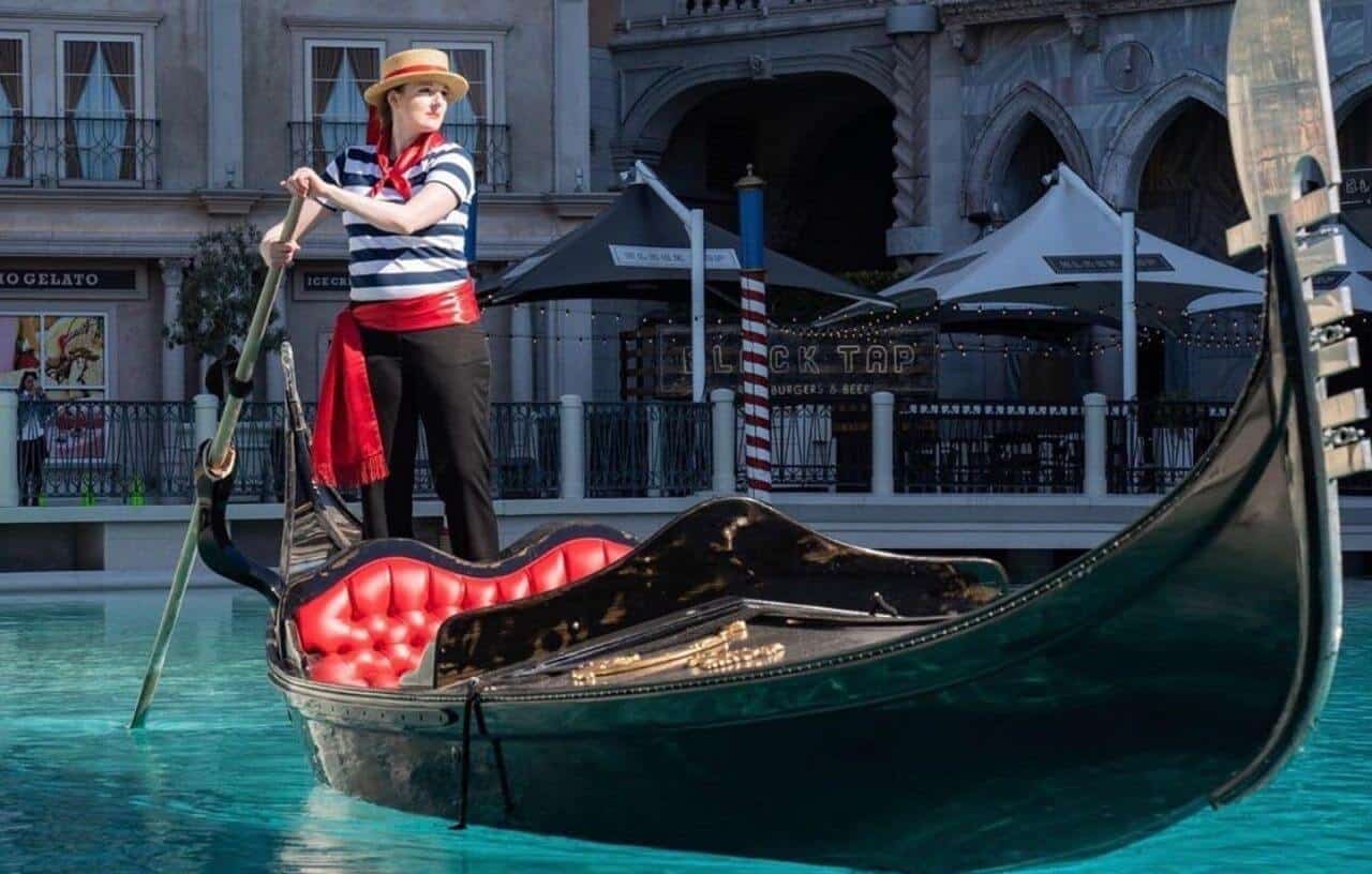 Woman in white and blue striped shirt and red bandana around her neck holding large stick pushing gondola on man made river in front of the Venetian resort in Las Vegas